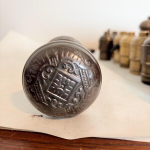 May include: A round, dark metallic door knob featuring an intricate embossed pattern with a central square motif surrounded by decorative scrollwork and geometric lines. The surface has a weathered, aged finish with visible dark patina throughout.
