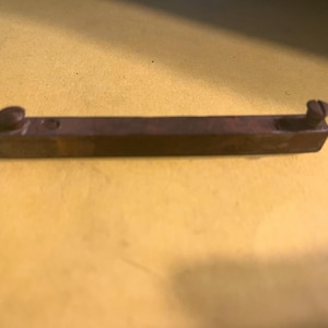 May include: A small, rectangular, brown metal object with two screws on each end. The object is lying on a yellow surface. The screws appear to be used to attach the object to something else.