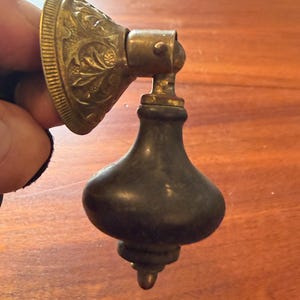 May include: Antique brass drawer pull with a decorative floral design on the backplate. The pull has a dark, bulbous body and a small, pointed base. The hardware is likely for a cabinet or drawer.
