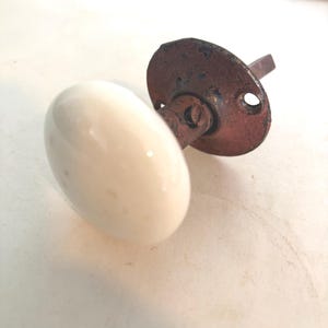 May include: A round white porcelain door knob featuring a weathered, dark metallic circular mounting plate with visible screw holes and a central square spindle shaft extending from the back, showing signs of age and oxidation on the metal.