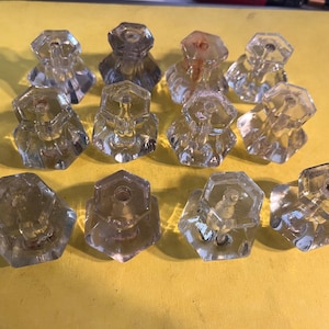 May include: A collection of twelve clear, faceted glass cabinet knobs. Each knob has a hexagonal shape and a screw post for attachment. The knobs are arranged on a yellow surface, showcasing their reflective qualities.
