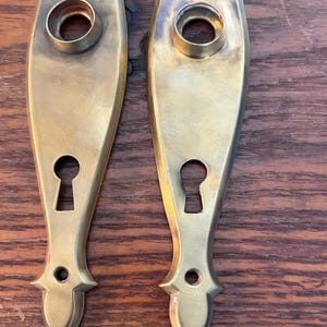 May include: Two antique brass door handle backplates. Each plate has an elongated shape with a keyhole, a circular opening for the handle, and decorative finials. The brass has a warm, aged patina, suggesting vintage craftsmanship.
