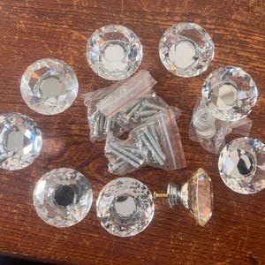 May include: A collection of clear, faceted crystal cabinet knobs with silver-tone hardware. The knobs are arranged on a wooden surface, along with screws and other hardware in small plastic bags. One knob has a gold-tone base.