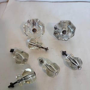 May include: A set of five clear glass cabinet knobs with an octagonal face and a fluted stem design. Each knob features a central metal screw mechanism with a threaded bolt and a small square nut attached for mounting to furniture or cabinetry.