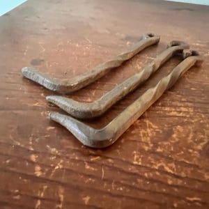 May include: Three weathered, L-shaped metal tools with a twisted design. The tools are a dark, rusty brown color and appear to be made of iron or steel. They are arranged on a wooden surface.