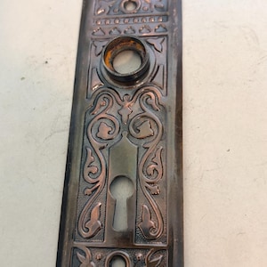 May include: A rectangular dark bronze door plate featuring an intricate embossed scrollwork pattern, a central keyhole, and a circular knob opening at the top. The plate includes two screw holes at the top and bottom with decorative border details.