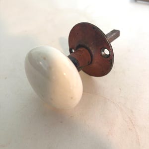 May include: A vintage door knob featuring a smooth, round white porcelain handle attached to a circular, weathered reddish-brown metal mounting plate with two screw holes and a square metal spindle extending from the back of the hardware.