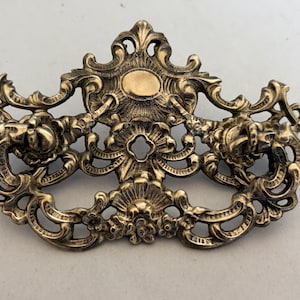 May include: A brass-colored metal drawer pull featuring an ornate, symmetrical design with intricate scrollwork, floral motifs, and a central oval plate. The hardware includes two swinging drop handles attached to the decorative backplate.
