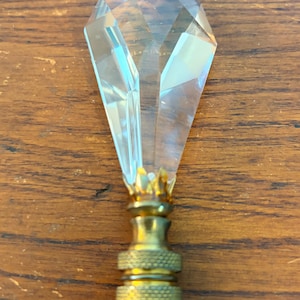 May include: A clear, faceted crystal lamp finial with a brass base. The crystal is diamond-shaped and tapers to a point. The brass base has a decorative, textured design.