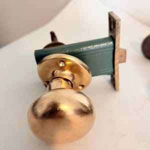 May include: A brass door knob with a spherical handle attached to a circular mounting plate. The mechanism features a rectangular, muted green metal housing with a brass faceplate and a protruding latch bolt, showing a metallic finish.