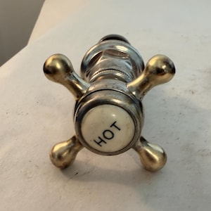 May include: A metal faucet handle with a cross-shaped design featuring four rounded arms. The center of the handle has a circular white ceramic insert with the word HOT printed in black text. The metal finish appears to be a polished brass or chrome.