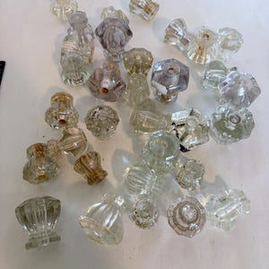 May include: A collection of clear glass cabinet knobs in various shapes and sizes, including faceted, fluted, and smooth designs. These hardware pieces feature central holes for mounting and exhibit transparent, reflective surfaces scattered across.