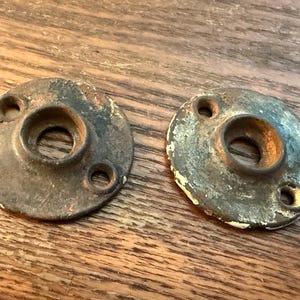 May include: Two antique, circular metal hardware pieces with a central hole and three smaller holes. The metal has a weathered, aged appearance with visible rust and paint loss, suggesting vintage use. The pieces are set on a wooden surface.