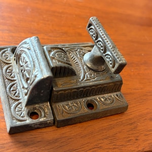 May include: An antique metal latch with ornate floral and geometric designs. The latch is a dark, aged bronze color and features a handle for easy opening and closing. This decorative hardware is suitable for cabinets or doors.