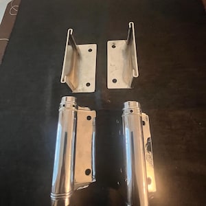 May include: Four metal boat hardware pieces. Two are rectangular with a curved top, and two are cylindrical with a rectangular base. All pieces are silver and appear to be made of stainless steel. The hardware is arranged on a dark surface.