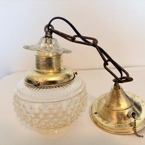May include: A hanging pendant lamp featuring a clear, hobnail-textured glass globe shade, a brass-toned metal cap, and a matching brass-toned ceiling canopy. The fixture is suspended by a dark metal chain and includes a pull-chain switch.