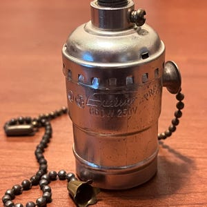 May include: A vintage silver-toned light socket with a pull chain and a decorative bell-shaped pull. The socket has the text "660 W 250V" and is set against a brown wooden surface. The pull chain is made of small, dark beads.