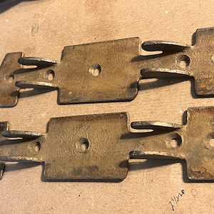 May include: Two vintage brass-toned metal latches. Each latch has a rectangular center with a hole and two curved extensions with holes. The latches are slightly tarnished, showing signs of age and use. They are likely for a cabinet or trunk.
