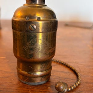 May include: Vintage brass light socket with a pull chain and a small, round pull. The socket has a cylindrical shape with a slightly wider base and a narrower top. The text "HARVEY" is visible on the side.