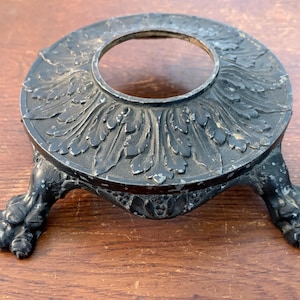 May include: Ornate, dark metal candle holder with a circular top and a central opening. The base features three claw feet. The top is decorated with a leaf pattern. The candle holder is on a wooden surface.