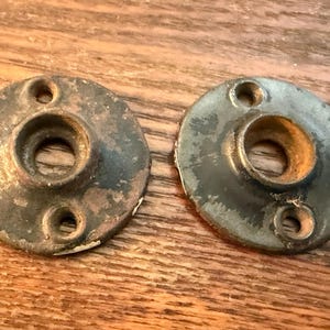 May include: Two antique, circular metal hardware pieces with a central hole and three smaller holes. The metal has a weathered, aged appearance with signs of rust and wear, suggesting they are vintage or salvaged items.