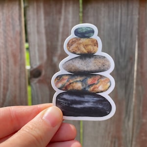 May include: A stack of five smooth, round stones in various shades of brown, gray, and black. The stones are stacked in a pyramid shape, with the smallest stone on top and the largest stone on the bottom.