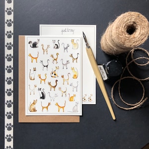 May include: A card with the text "Just to say" printed on the front. The card features a pattern of 30 illustrated cats in various poses and colours. The card is on a brown background.