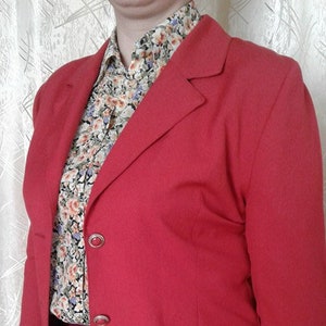 May include: A red blazer with a single-breasted closure and three buttons. The blazer has a pocket on the right side and a floral patterned shirt is visible underneath.