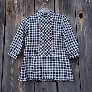 May include: A black and white gingham dress with a pleated skirt and orange buttons. The dress has a high neck and long sleeves.