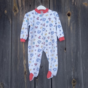 May include: White baby sleeper with a colorful pattern of teddy bears, bows, and stars. The sleeper has long sleeves, a zipper down the front, and red trim on the collar, cuffs, and feet. The garment is hanging on a white hanger.