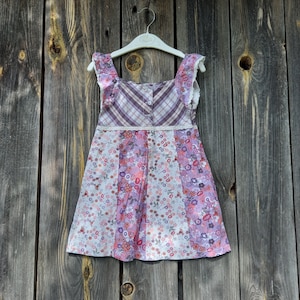 Puede incluir: Un vestido floral blanco y morado con un corpiño a cuadros y ribete de encaje. El vestido está colgado en una percha blanca sobre un fondo de madera.