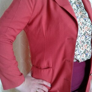 May include: A red blazer with a single button closure and a pocket on the left side. The blazer is worn over a floral patterned blouse and a burgundy skirt.