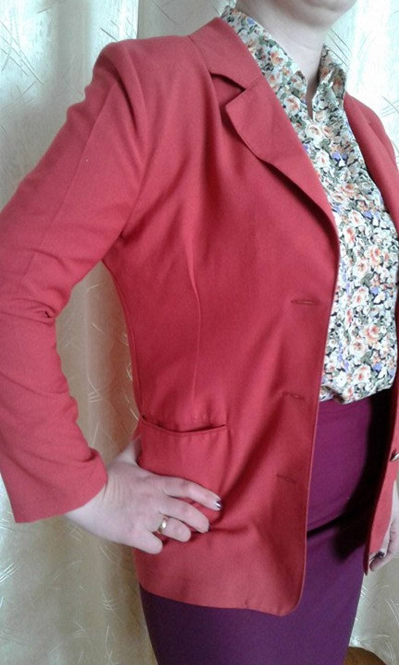 May include: A red blazer with a single button closure and a pocket on the left side. The blazer is worn over a floral patterned blouse and a burgundy skirt.
