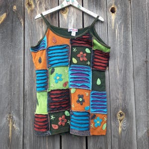 Könnte beinhalten: Ein farbenfrohes Patchwork-Tanktop in Grün, Orange, Blau und Rot. Das Oberteil ist mit verschiedenen gestickten Blumen und Blättern verziert. Das Etikett auf dem Oberteil lautet "Earth Dolls".