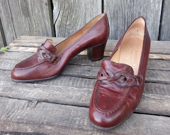 May include: A pair of brown leather heeled shoes with a braided detail on the front. The shoes have a low block heel and a pointed toe.