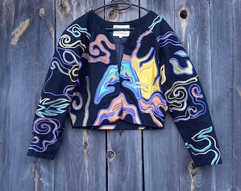 Puede incluir: Una chaqueta negra corta con mangas largas y un estampado abstracto colorido. El estampado presenta remolinos y líneas en tonos de azul, verde, amarillo, naranja y morado. La chaqueta tiene cuello en V y cierre de botón.