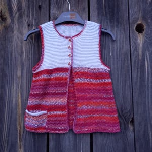 May include: A sleeveless knitted vest with a white top section and a striped red, pink, and orange bottom section. The vest has a button closure and a small pocket on the left side.