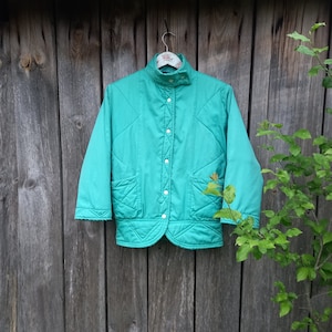 May include: A teal quilted jacket with a button-up front and a stand-up collar. The jacket is hanging on a wooden hanger against a wood panel background.