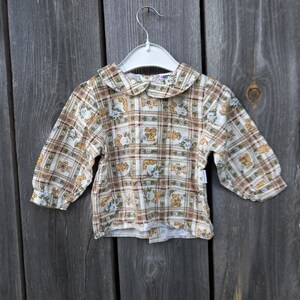 Puede incluir: Una camisa blanca de manga larga con un estampado de cuadros marrones y blancos y un pequeño estampado de osos. La camisa tiene un cuello Peter Pan.