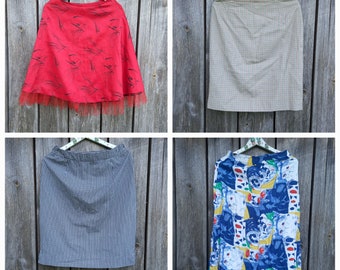 May include: Four different skirts hanging on hangers. The first skirt is red with a black floral pattern and a black tulle trim. The second skirt is a beige and brown plaid. The third skirt is black and white gingham. The fourth skirt is white with a blue, yellow, and red abstract pattern.