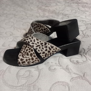 May include: A pair of black and white leopard print sandals with wooden heels. The sandals have a criss-cross strap design.
