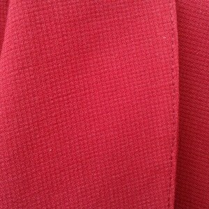 May include: Close-up of a red blazer with a textured fabric. The blazer has a single button closure and a notched lapel.