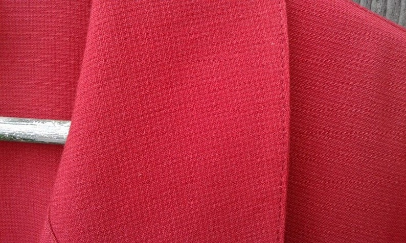 May include: Close-up of a red blazer with a textured fabric. The blazer has a single button closure and a notched lapel.