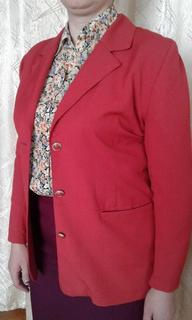 May include: A red blazer with a single-breasted closure and three buttons. The blazer has a pocket on the right side and a floral patterned shirt is visible underneath.