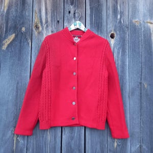 Puede incluir: Un cárdigan rojo de punto con cierre de botones y detalles de punto de cable en las mangas. El cárdigan está colgado en una percha de madera.