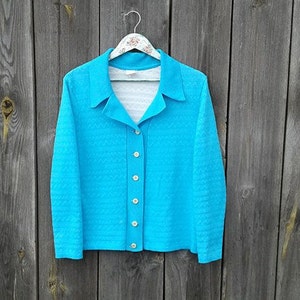 May include: A turquoise blue cardigan with a button-up front and a collar. The cardigan is made of a textured fabric and has long sleeves.