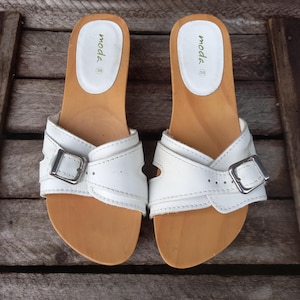 Pode incluir: Um par de sandálias de couro branco com solas de madeira. As sandálias apresentam uma tira de couro branco com uma fivela prateada. A palmilha é castanho-clara e as sandálias estão rotuladas "moda", tamanho 38.