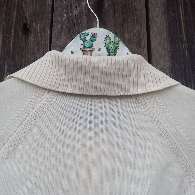 Puede incluir: Un su&eacute;ter de punto blanco con cuello de canal&eacute;. El su&eacute;ter est&aacute; colgado en una percha con un dise&ntilde;o de cactus pintado.
