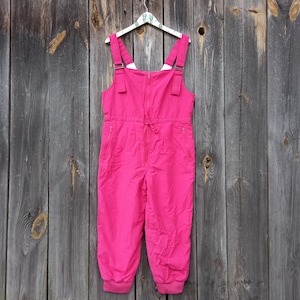May include: Pink ski overalls with a zipper closure and adjustable straps. The overalls have two side pockets and a drawstring at the waist.