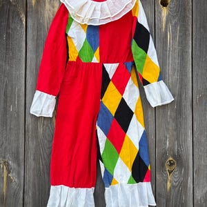 Puede incluir: Un disfraz de payaso vintage con corpiño y pantalón rojos, y un colorido estampado de diamantes en un lado. El disfraz presenta un cuello, puños y dobladillo con volantes blancos. El atuendo de manga larga cuelga de una percha blanca sobre un fondo de madera desgastada.